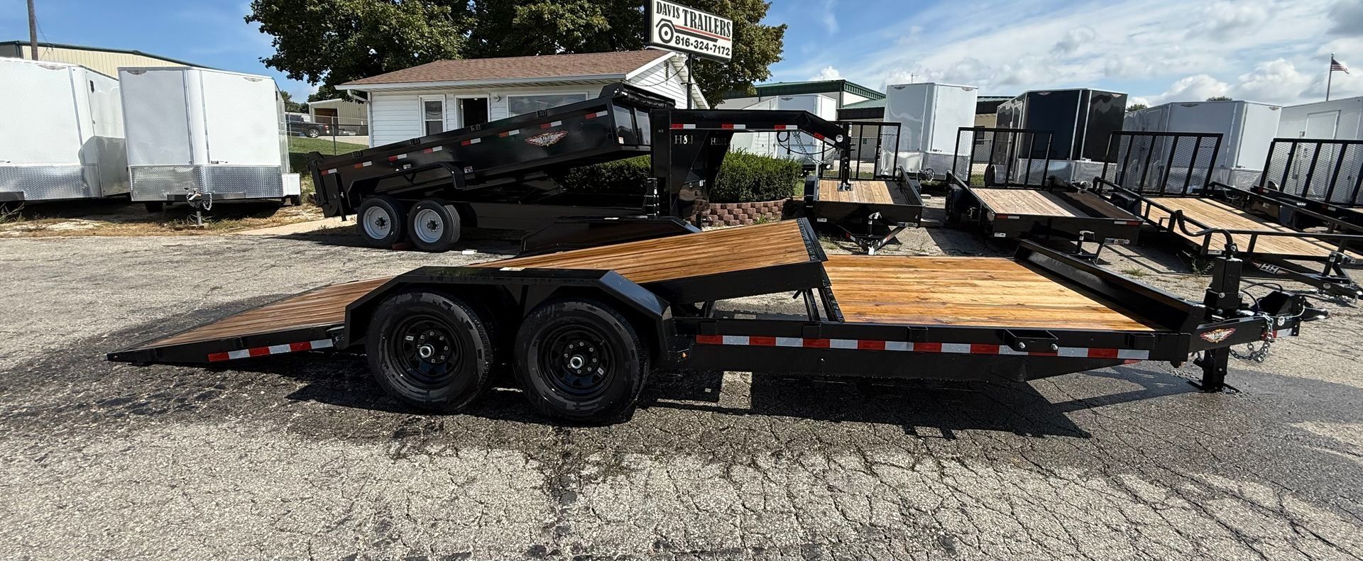 A flatbed trailer with wooden deck sits on gravel. Other trailers and buildings are in the background under a blue sky.