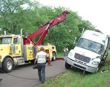 A yellow tow truck is positioned on an asphalt road to pull a white semi-truck out of a grassy ditch.