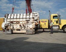 A large white industrial engine being lifted by a crane next to a yellow semi-truck on a concrete lot.