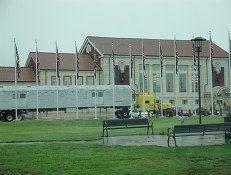 A large train car sits in front of a tan building with many flags flying in a grassy yard under a gray sky.