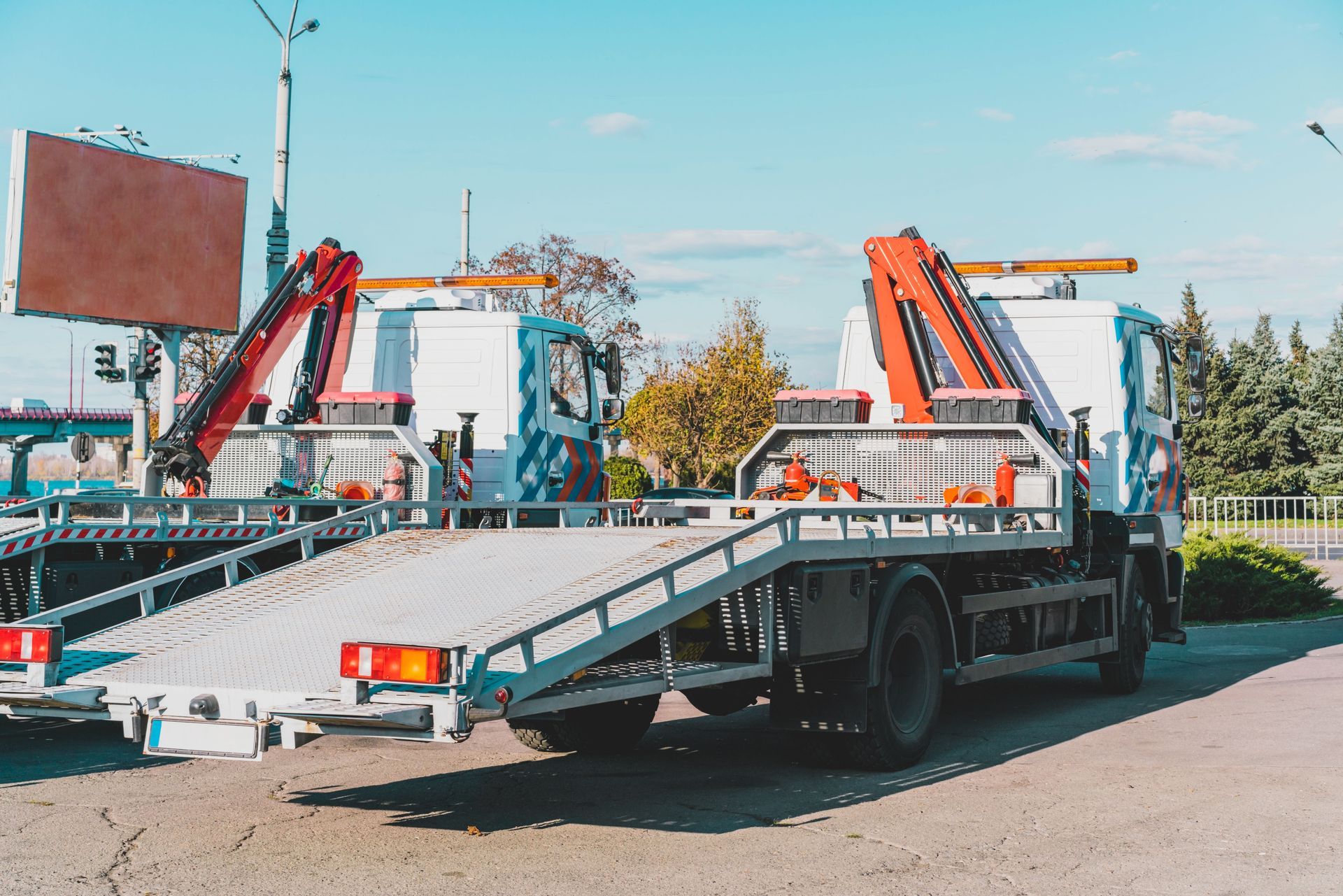 Two white flatbed tow trucks with orange hydraulic cranes parked side-by-side on an asphalt lot under a blue sky.