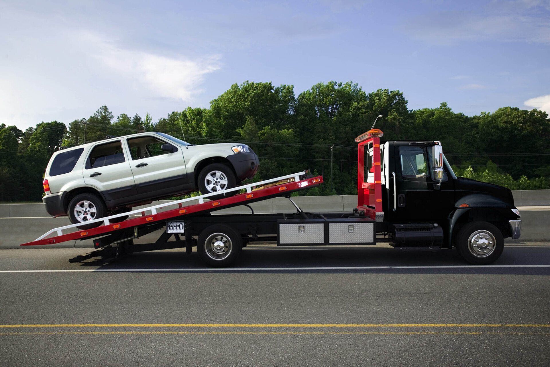 A flatbed tow truck carrying a light-colored SUV along a paved road with trees in the background.