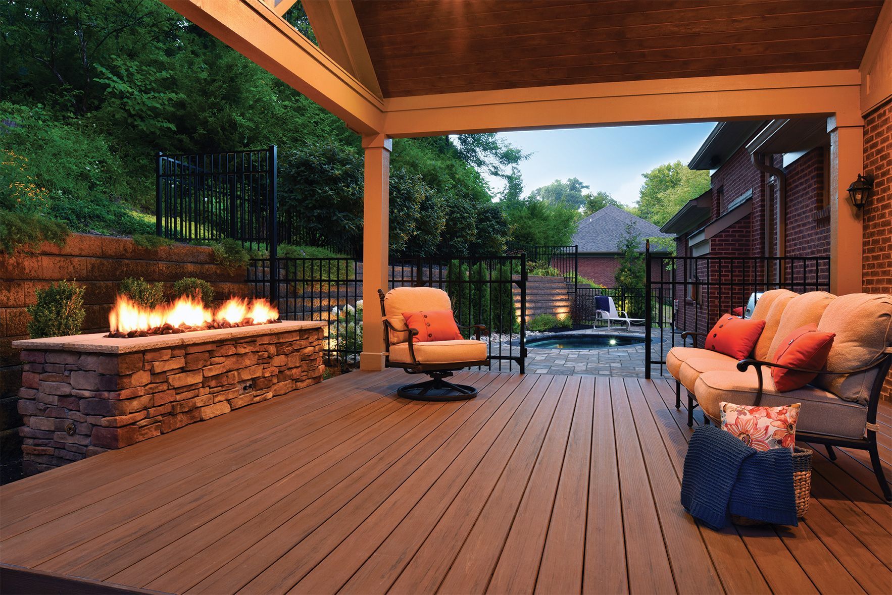 Covered wooden patio with lounge chairs, fire pit, and warm evening lighting overlooking a garden and pool area