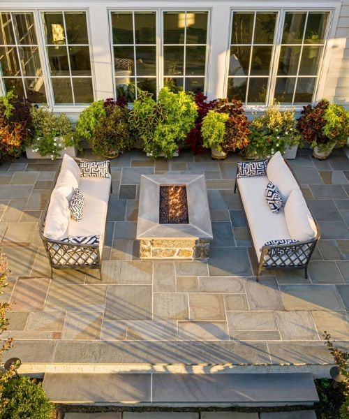 Patio seating area with white couches, fire pit, and potted plants in front of house windows