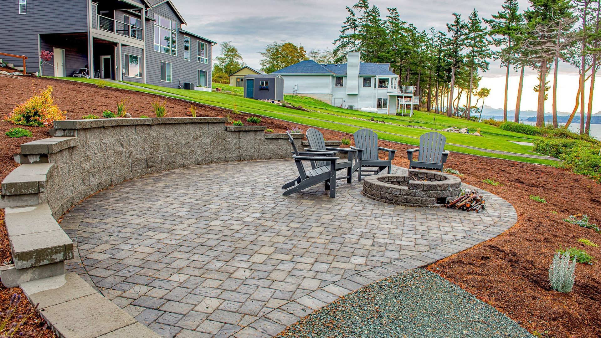 Patio with stone pavers, fire pit, chairs, and landscaped backyard overlooking trees and water