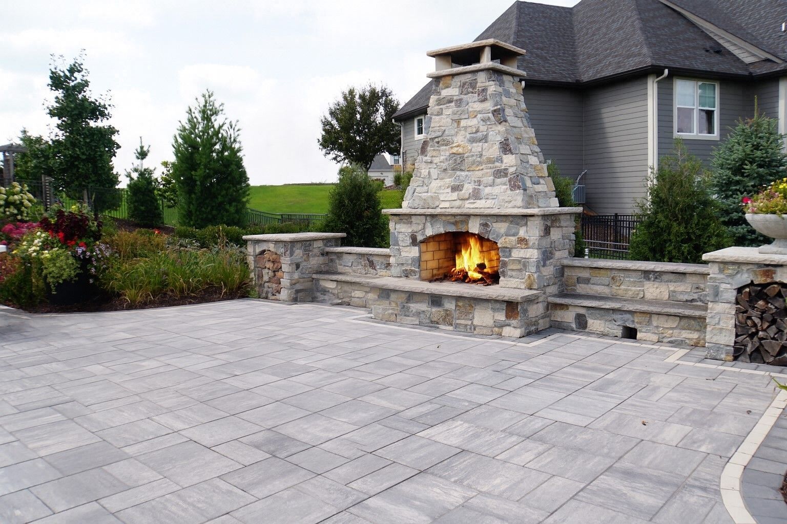 Stone outdoor fireplace lit on a paved patio beside a gray house and landscaped garden.