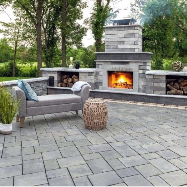 Outdoor patio with stone pavers, built-in fireplace, gray sectional sofa, and wicker stool surrounded by trees
