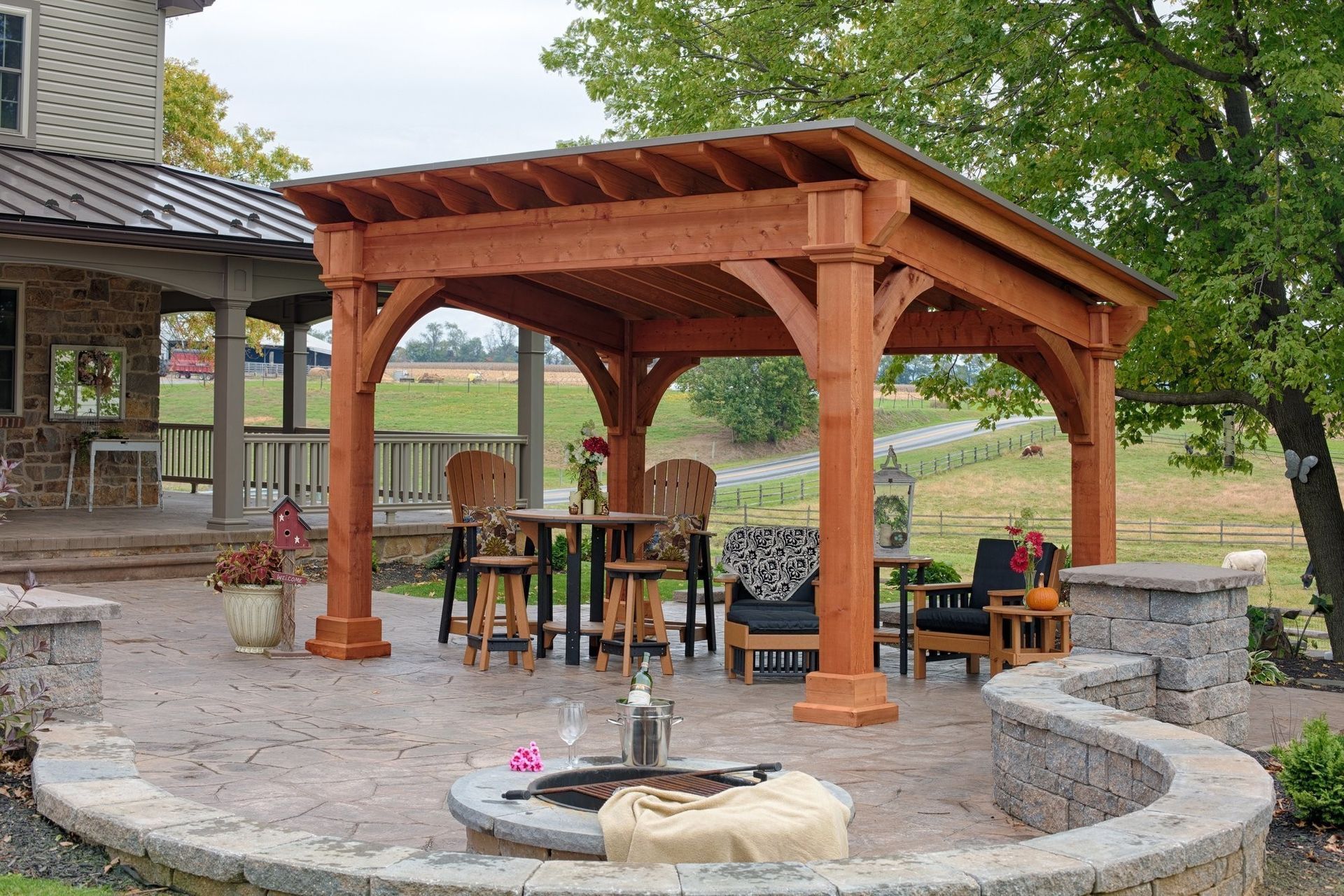 Wooden pergola shelter with picnic tables in a stone courtyard beside a fire pit