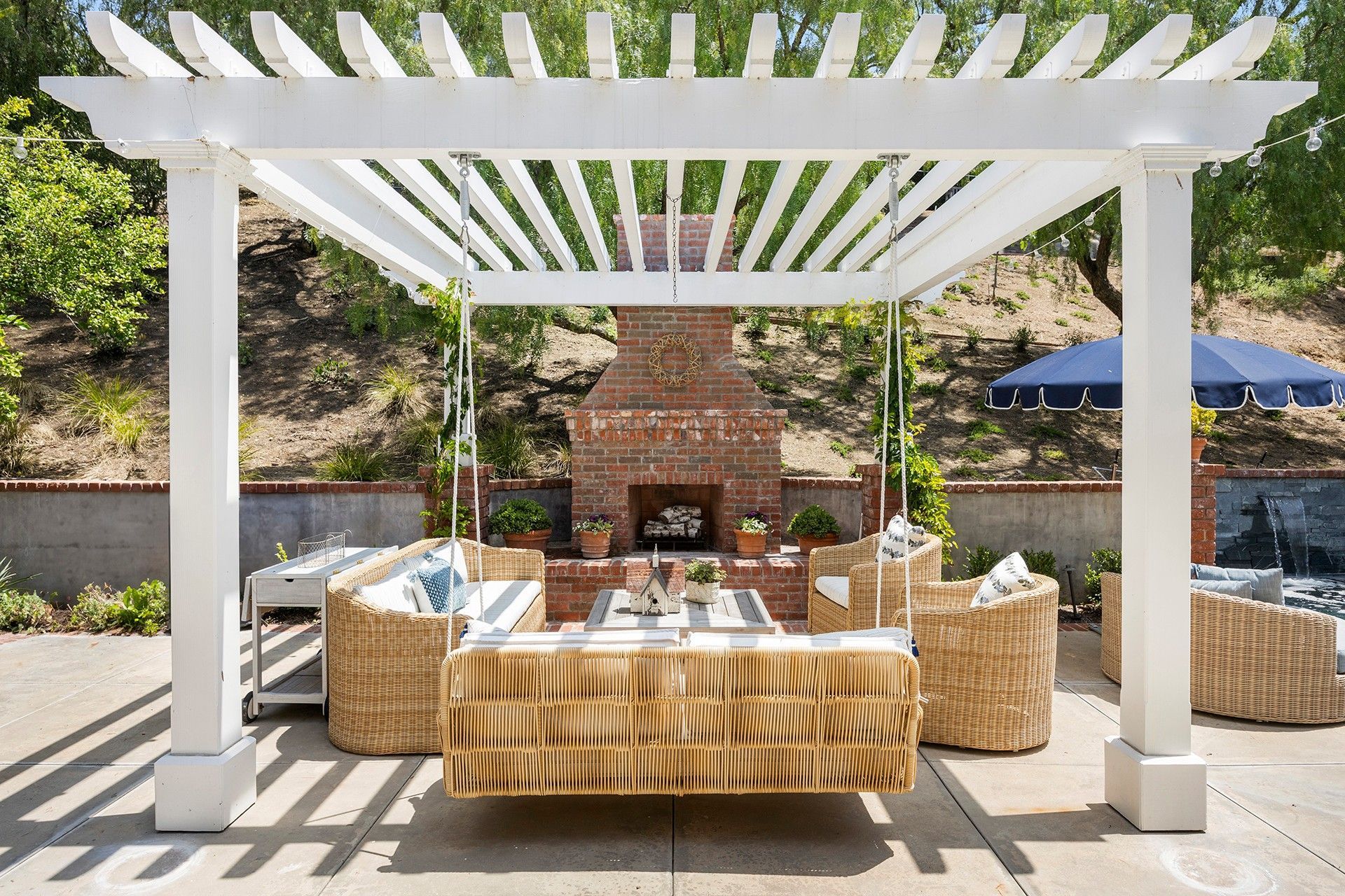 Outdoor patio dining area under a white pergola with wicker furniture and a brick fireplace