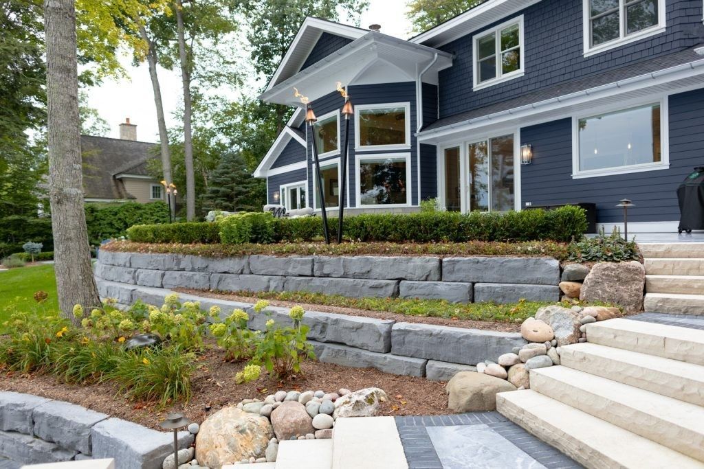 Front yard with stone retaining walls and steps leading to a gray house with a bay window and landscaped garden.