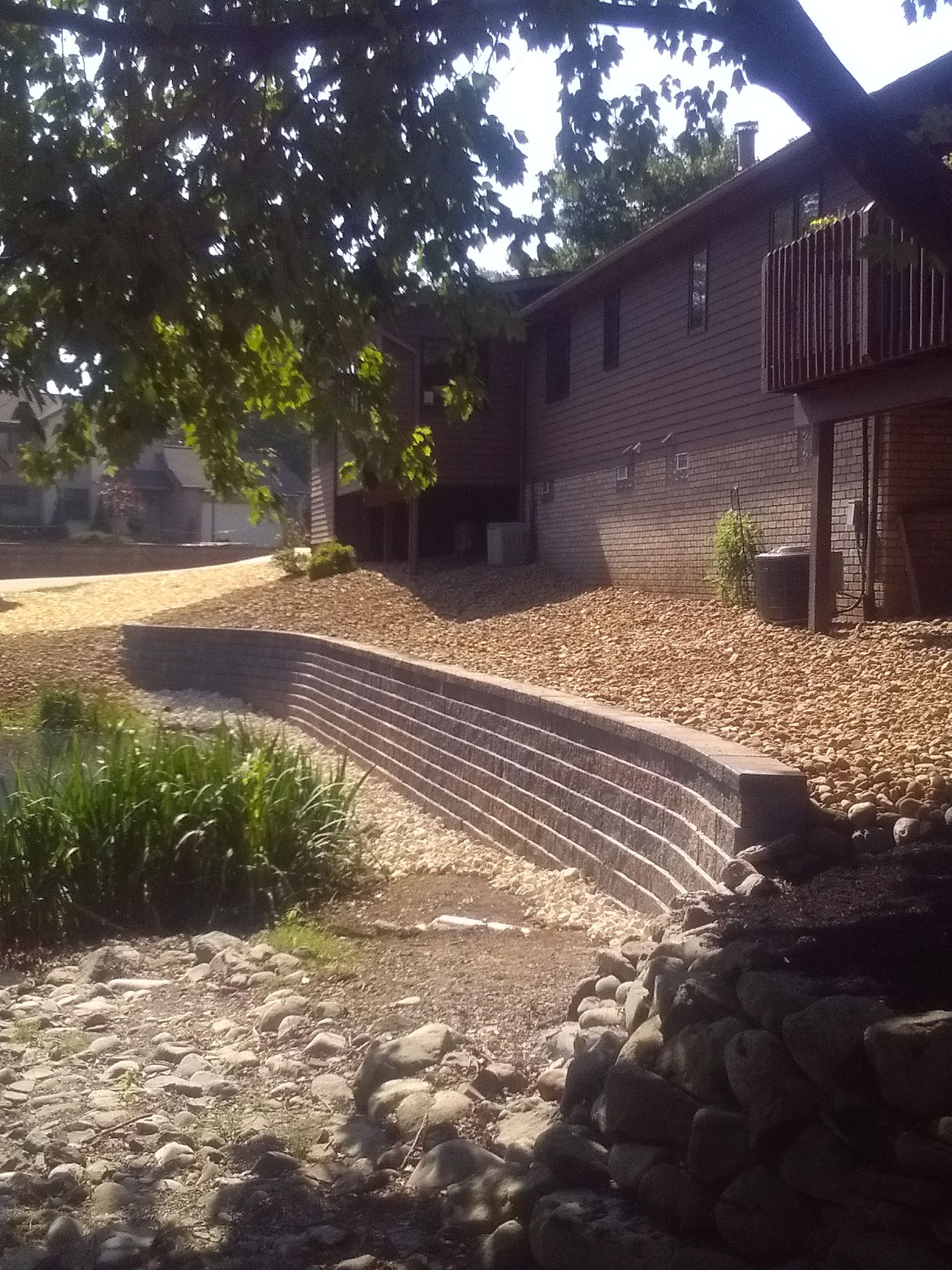 Curved stone retaining wall beside a brick building and leafy trees in a landscaped yard
