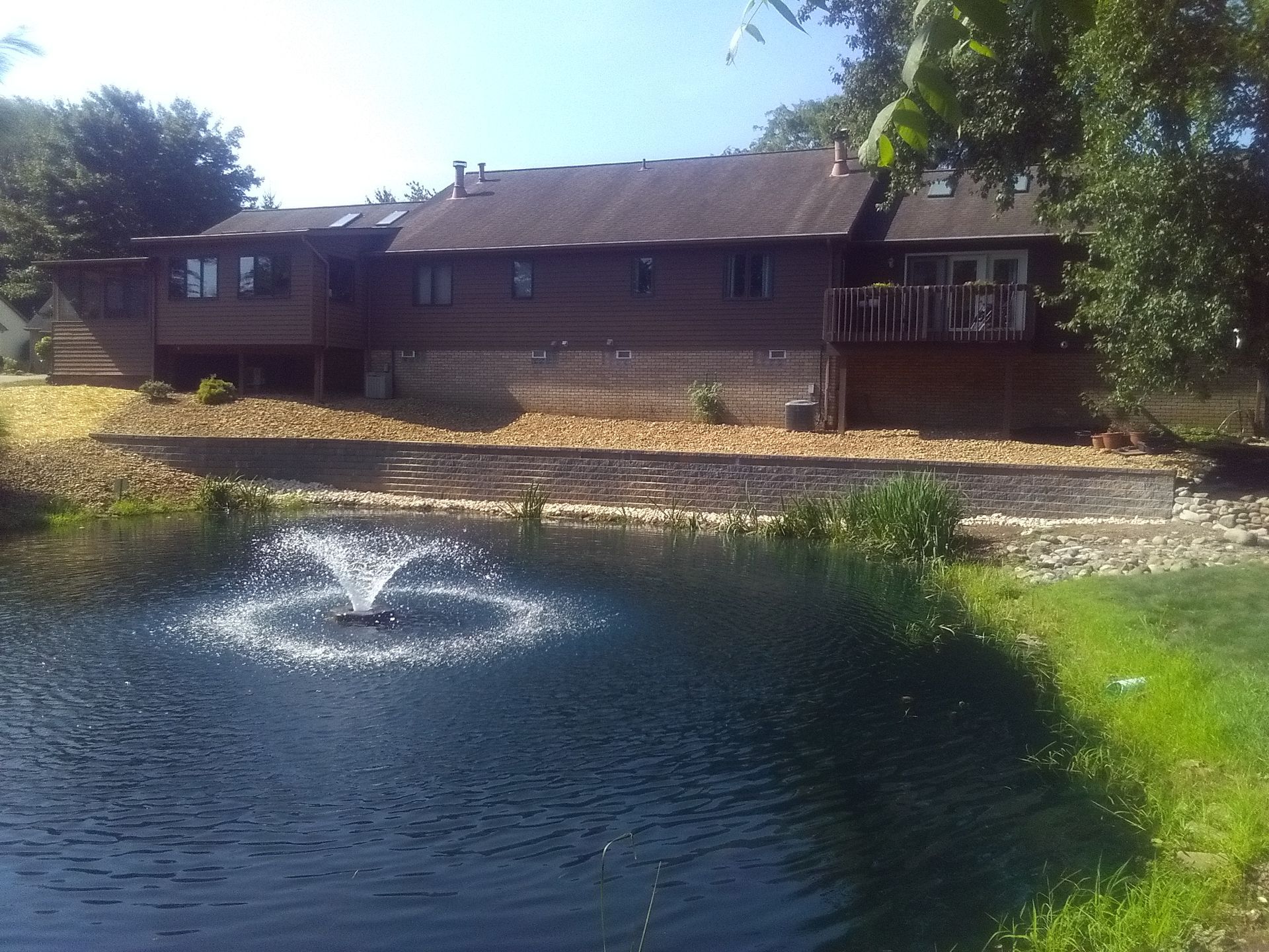 Brick house beside a pond with a fountain, under trees on a sunny day