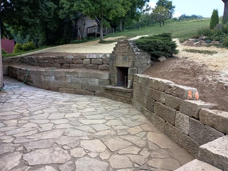 Stone patio with curved retaining walls and a small built-in fireplace in a landscaped yard