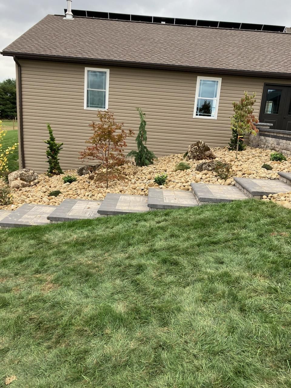 Side yard with tan house siding, small windows, stepping stones, and a landscaped bed with shrubs and mulch.