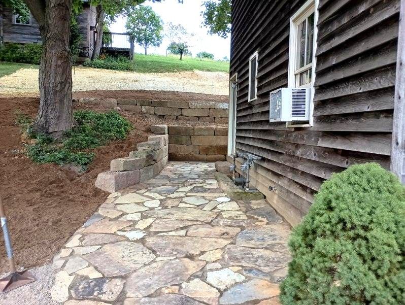 Stone walkway beside a wooden house, with steps up to a dirt yard and a tree nearby