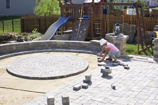 Child in a white hat builds with gray blocks on a brick patio beside a playground slide.