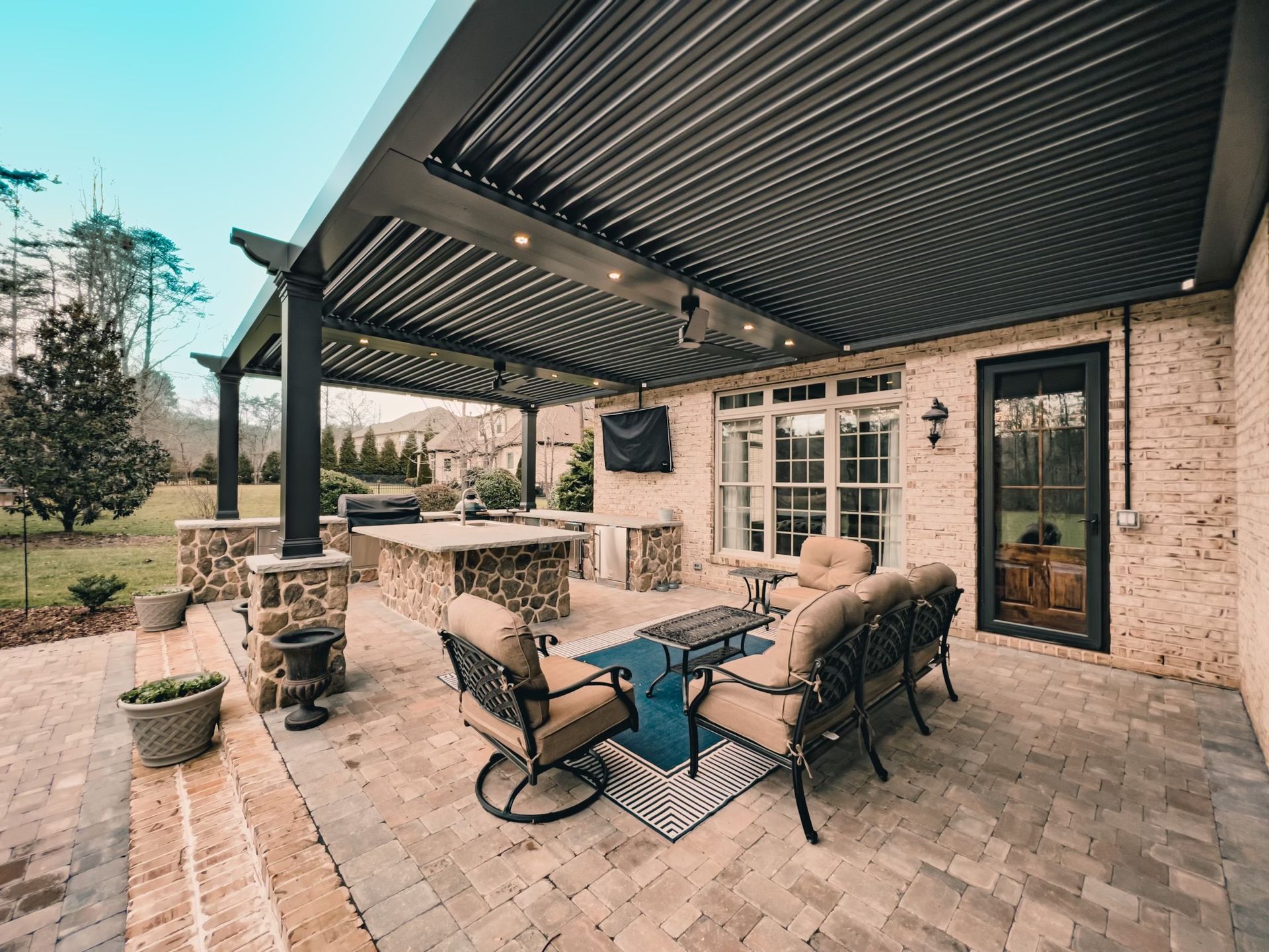 A patio with a pergola , couch , chairs , table and television.
