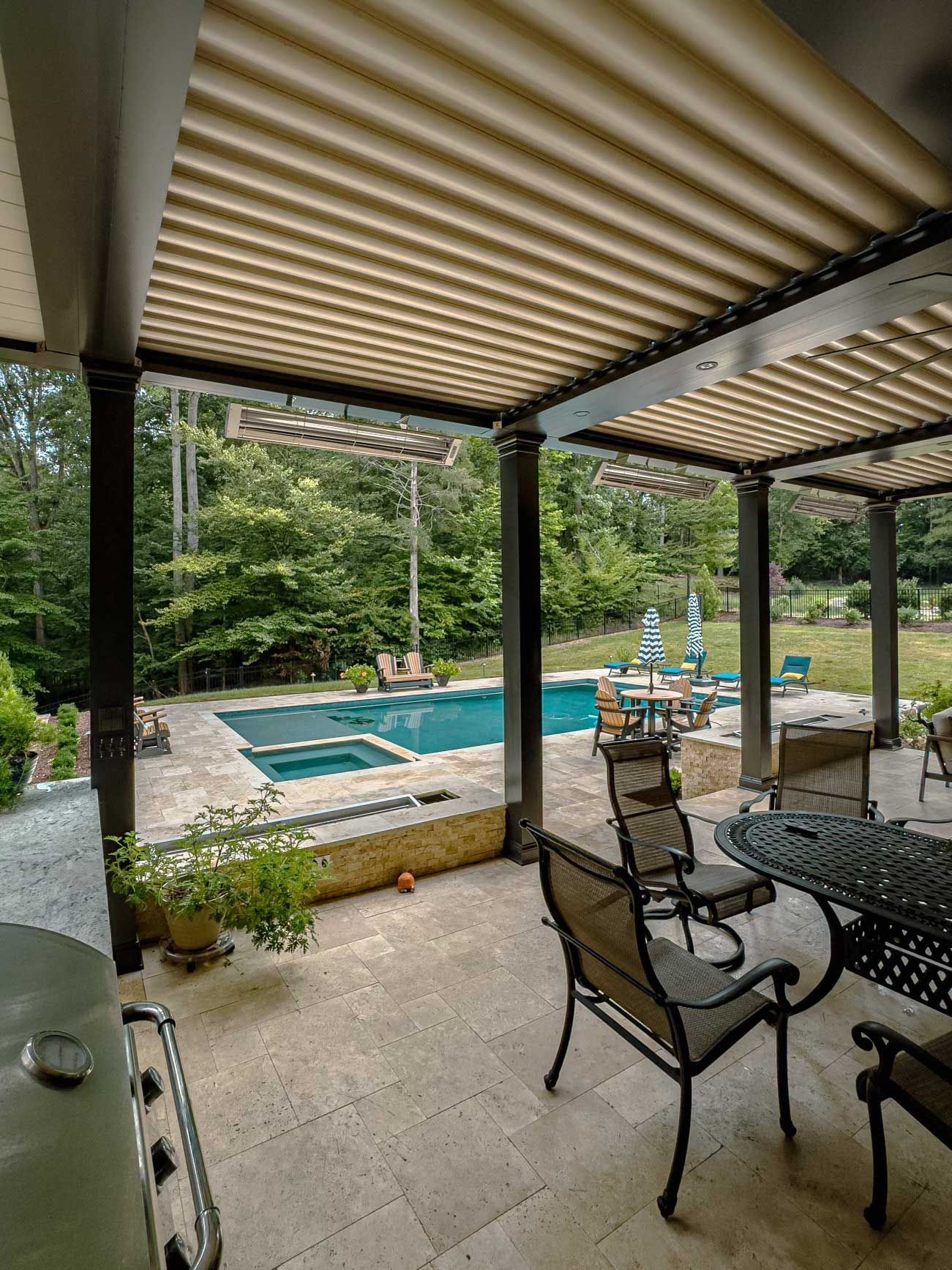 A patio with a table and chairs under a pergola with a pool in the background