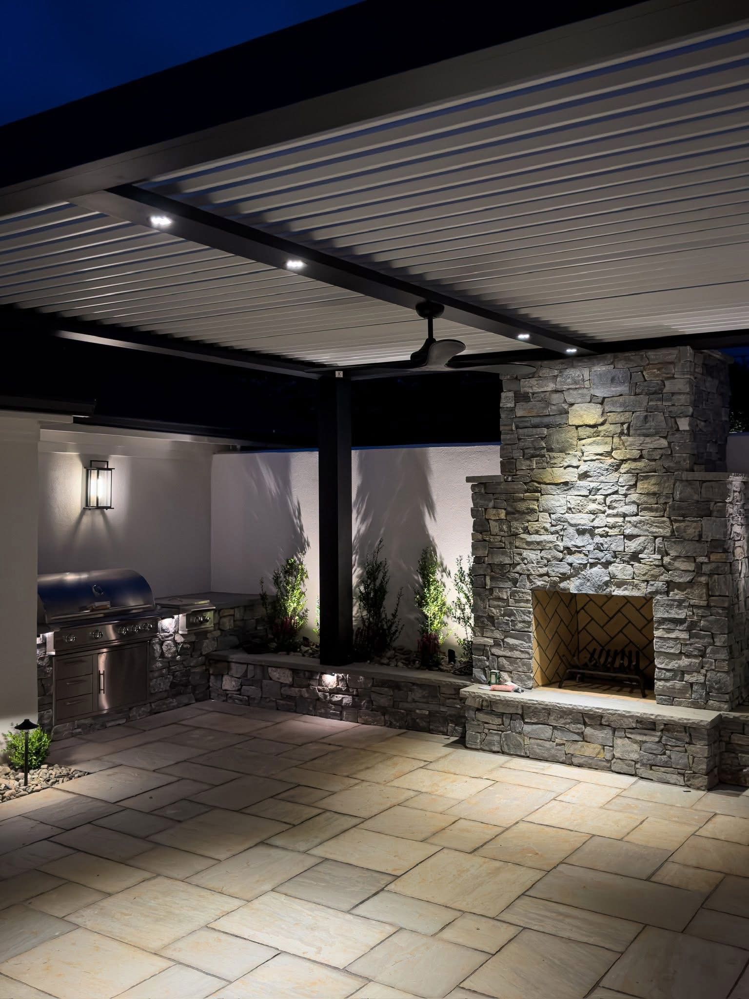 A patio with a fireplace and a ceiling fan at night.