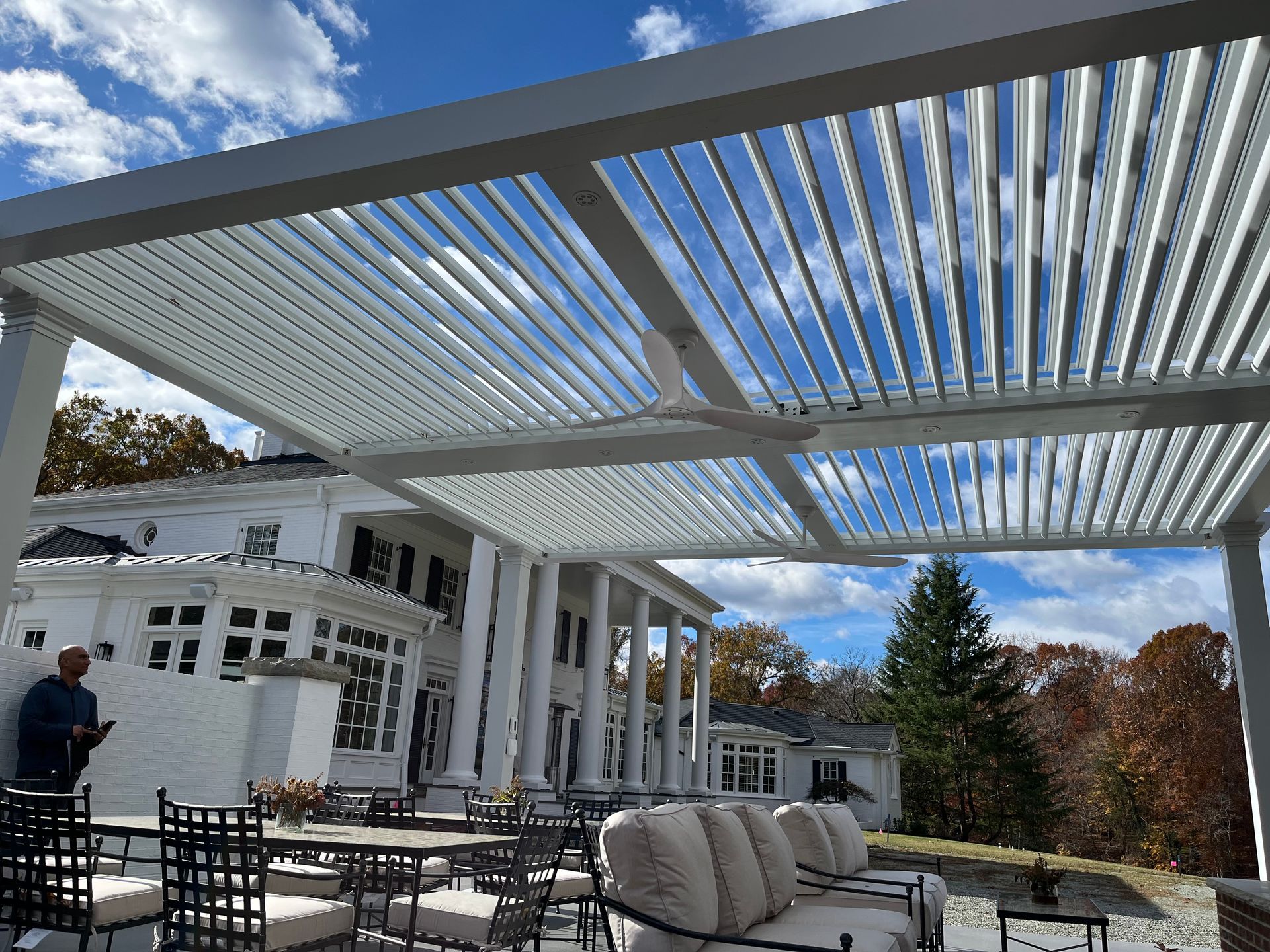 A man is standing under a pergola in front of a large white house.