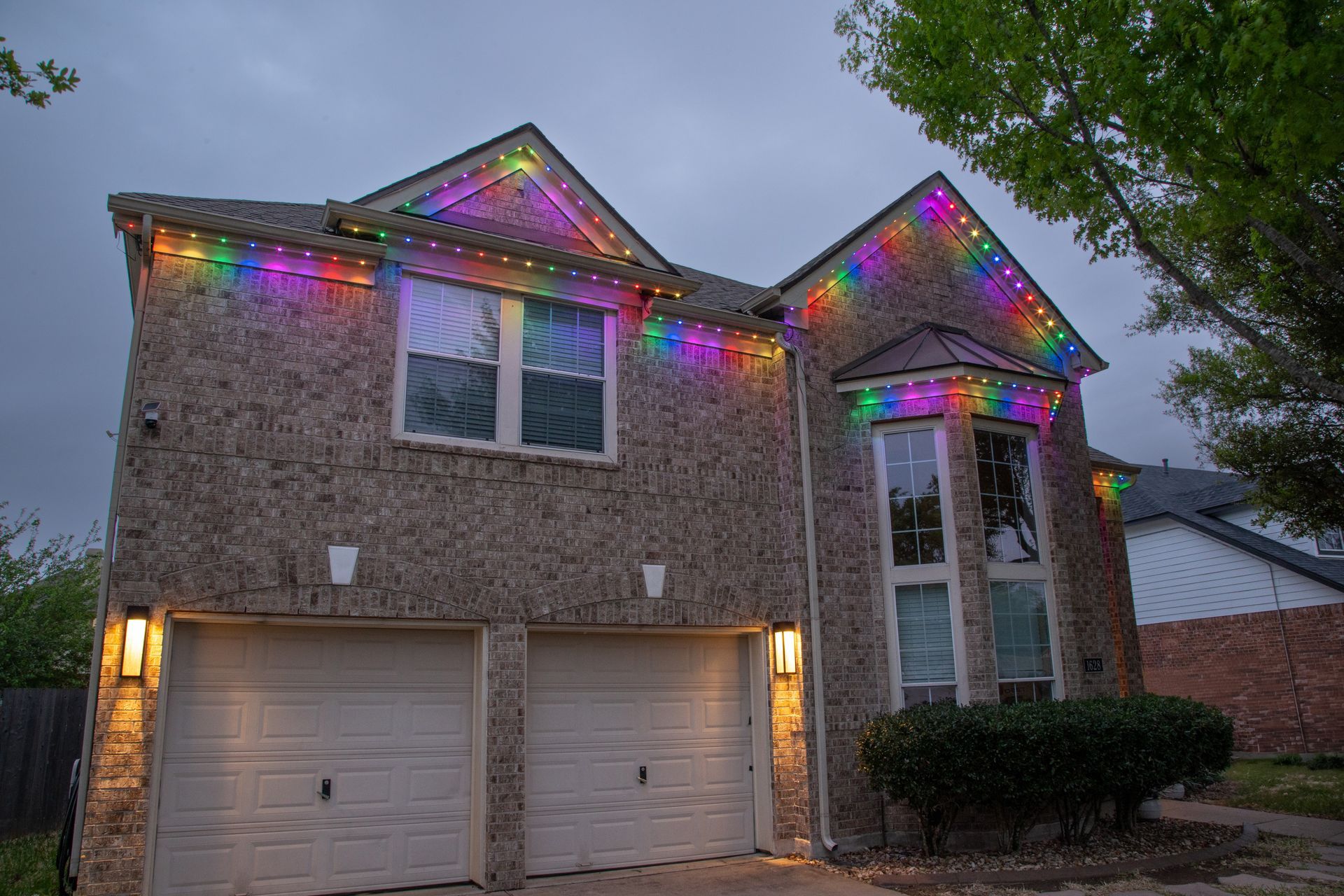 A large brick house is decorated with rainbow colored Christmas lights.