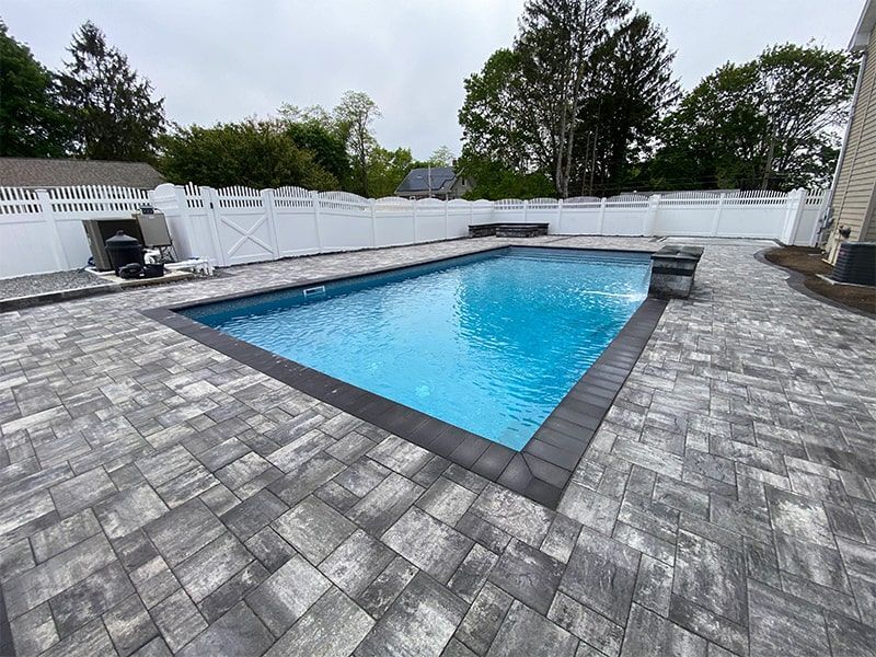A large swimming pool is surrounded by a brick patio and a white fence.
