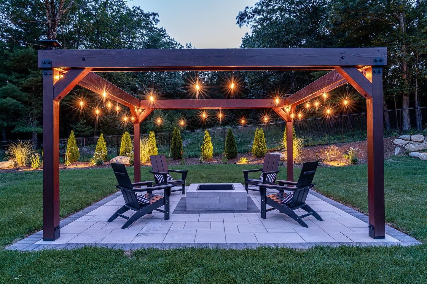 A cozy backyard patio with a wooden pergola strung with lights, surrounding a fire pit and lounge chairs.