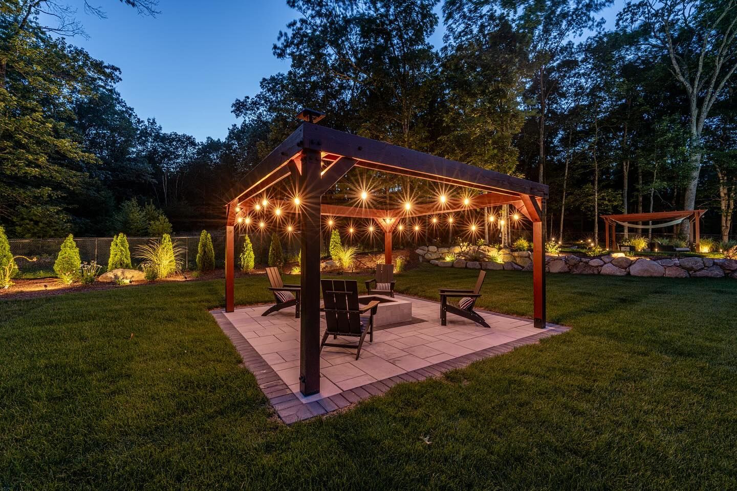 A lit pergola with string lights over a fire pit with chairs, in a backyard at dusk.