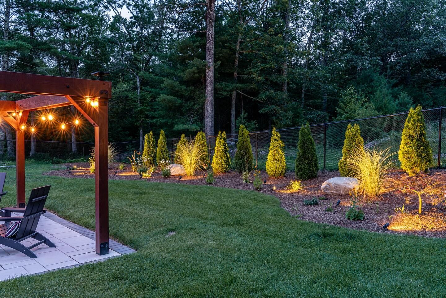 A backyard scene at dusk, with a pergola lit by string lights, a grassy lawn, and landscaping with up-lit trees.
