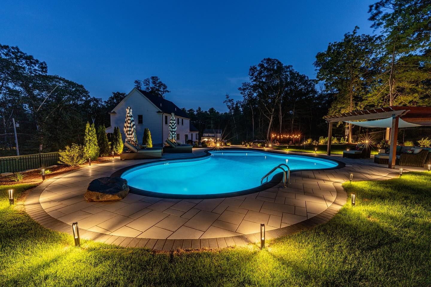 Night view of a backyard pool lit with blue water and pathway lights, a house in the background, trees and a pergola.