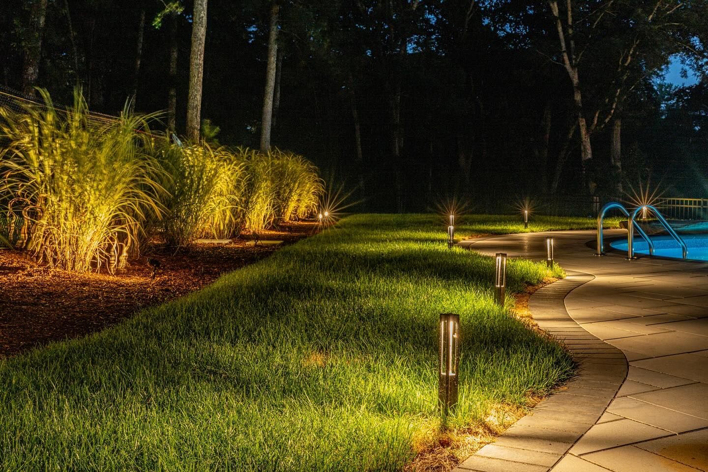 Pathway and landscaping illuminated at night with ground lights beside a pool in a dark forest.