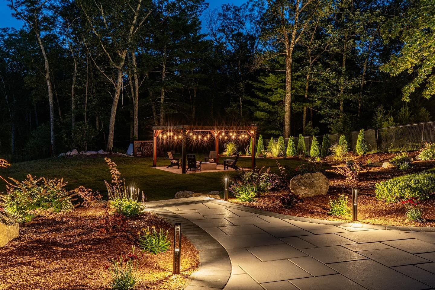 A backyard at night, lit by pathway lights leading to a pergola with string lights. Trees surround the seating area.