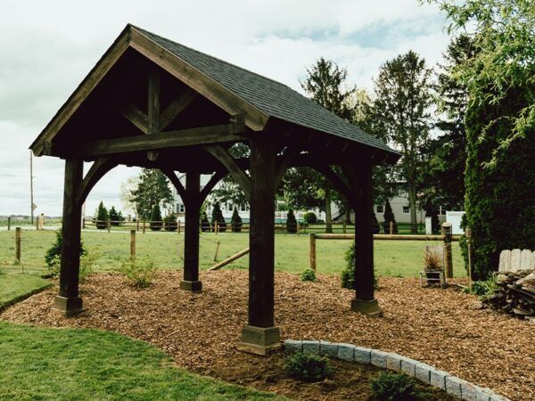 A wooden pavilion with a black roof sits in the middle of a grassy field