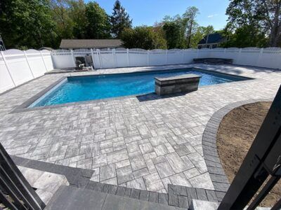 A large swimming pool is surrounded by a brick patio and a white fence.