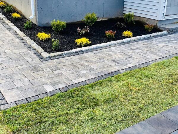 A brick walkway leading to a house with a garden in the corner.