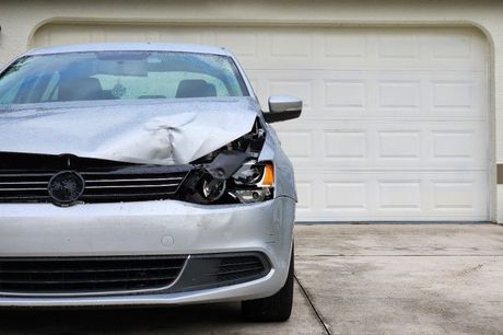 A silver car with a damaged headlight is parked in front of a garage door.