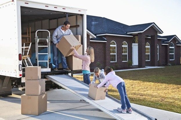 People unloading moving boxes from a truck via a ramp outside a suburban house