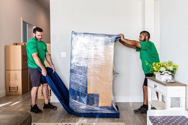 Two movers wrapping a large mirror in blue plastic inside a bright living room