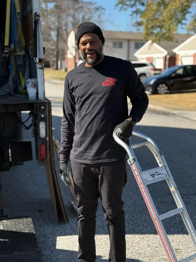 Man in black clothes and beanie smiling beside a truck, holding a metal ladder outdoors on a sunny day