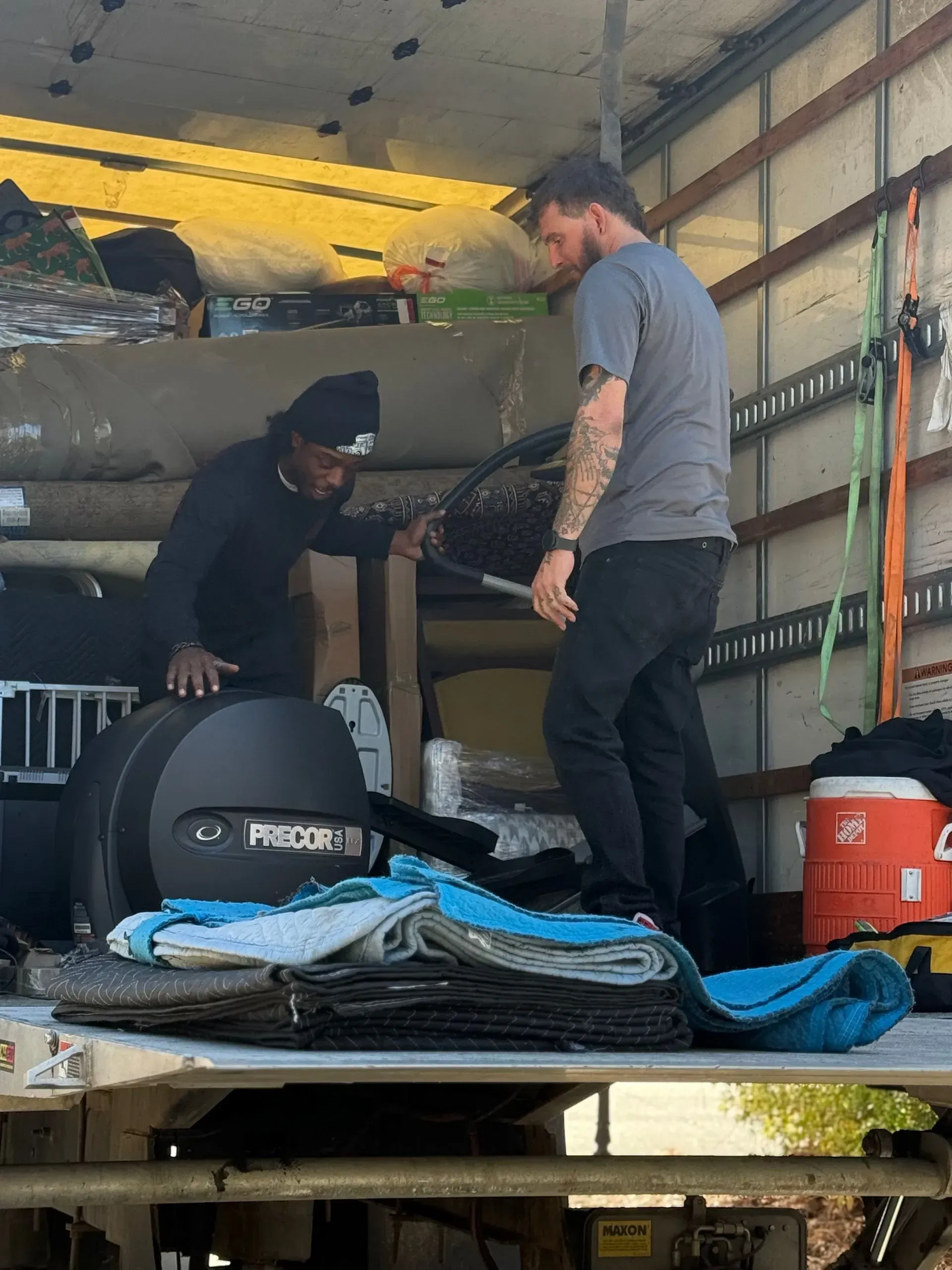 Two people unloading stacked cargo inside a truck, with bags, straps, and covered equipment around them.