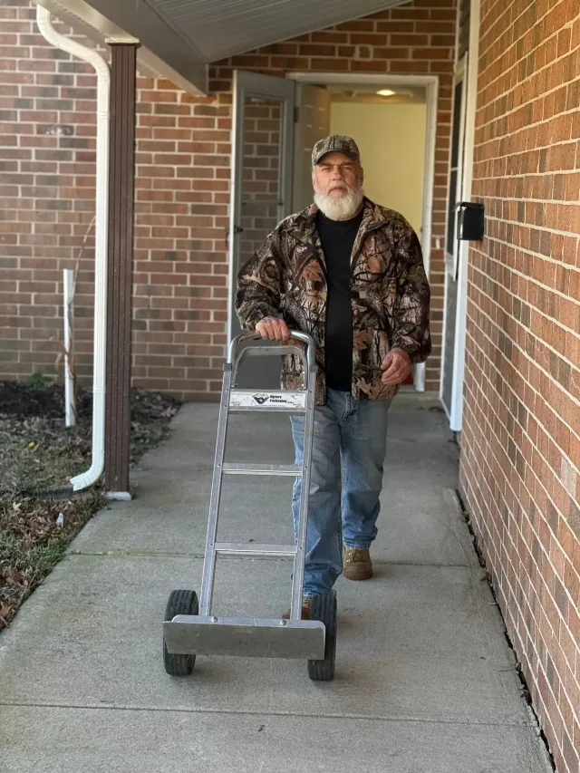 Man standing on a front porch beside a wheeled walker, wearing a camo jacket and cap.
