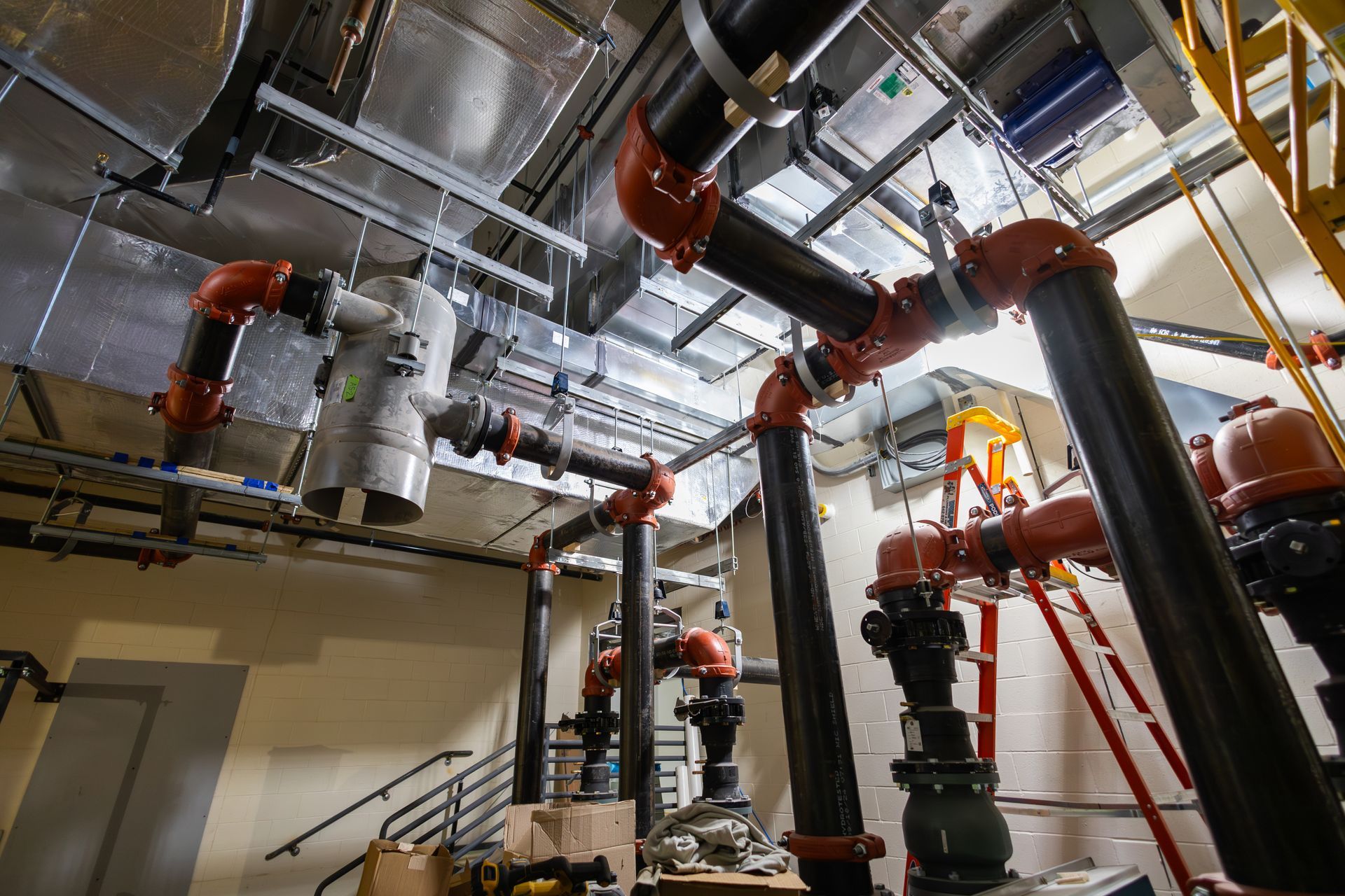 Pipes in a mechanical room with red elbows and black pipes. A set of stairs and ladders are visible.