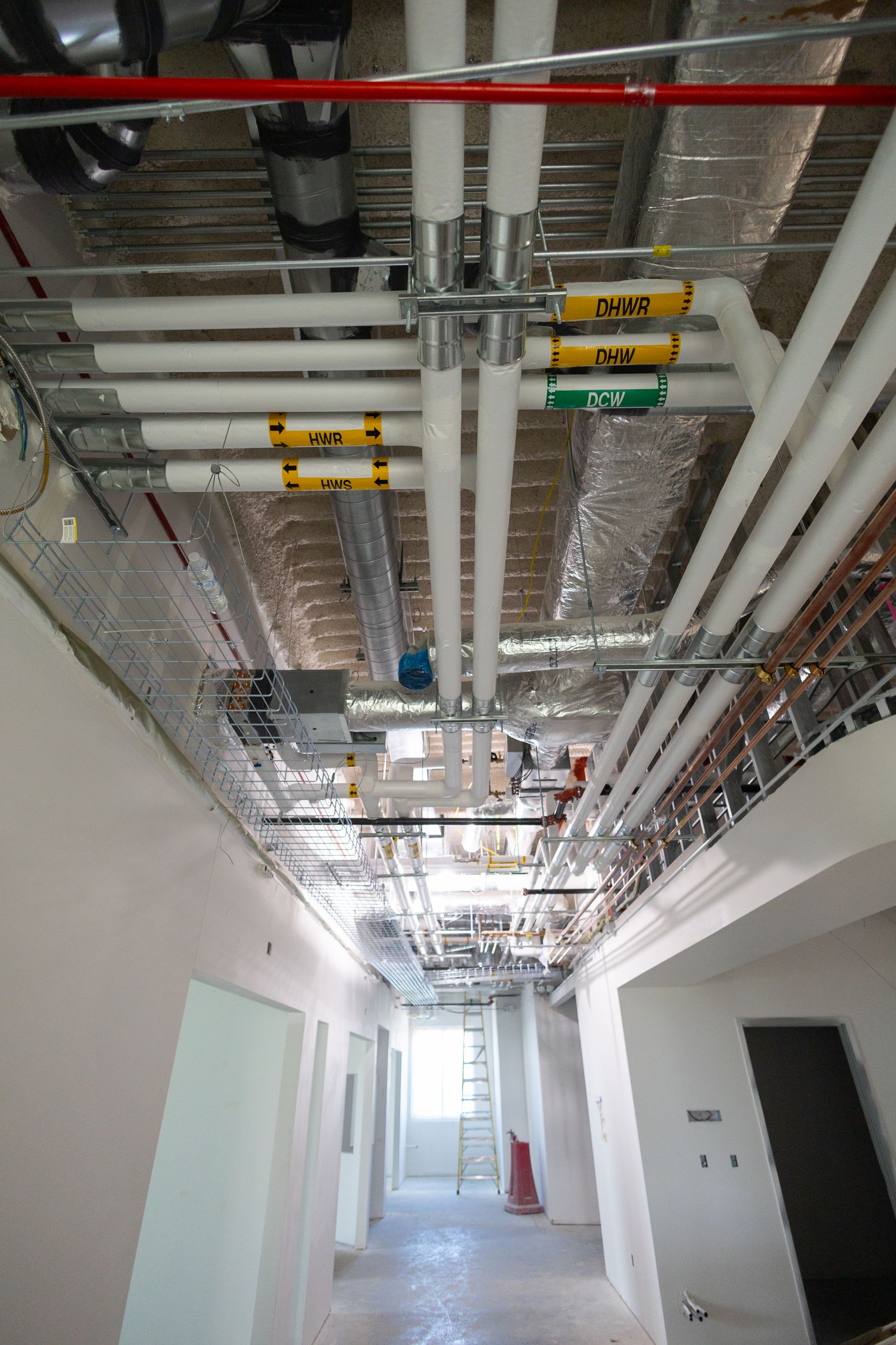 Construction hallway with overhead pipes, ductwork, and exposed ceiling.