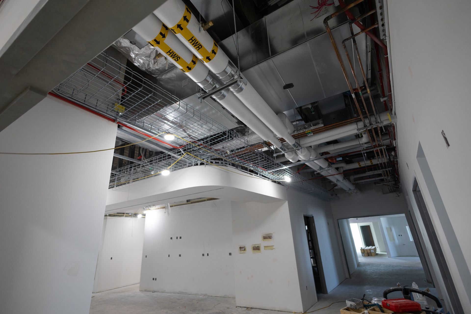 Interior construction site with exposed pipes and wiring on the ceiling, white walls, and unfinished floors.