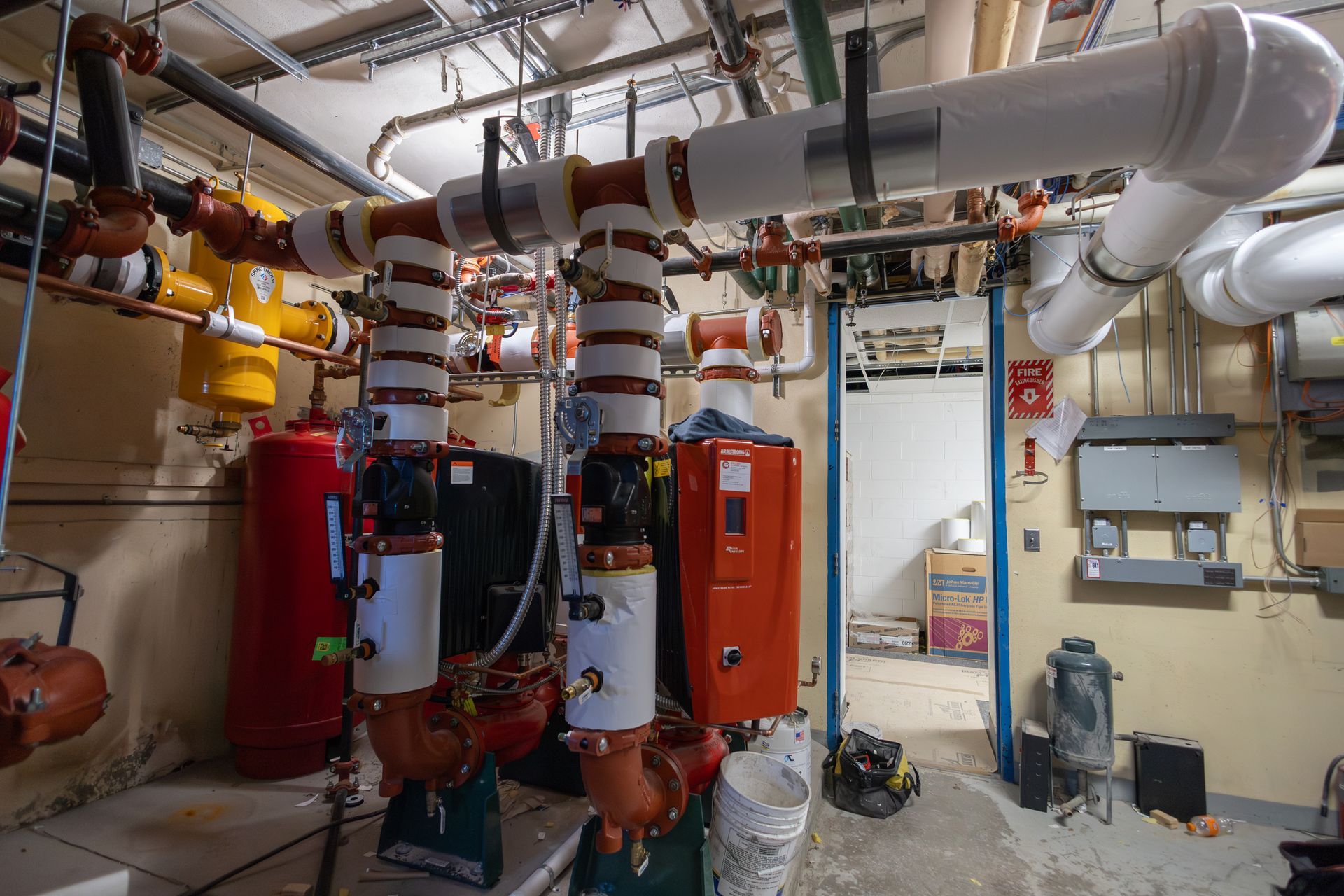 Plumbing and heating equipment in an industrial room; red, yellow, and white pipes, boilers, and tanks.