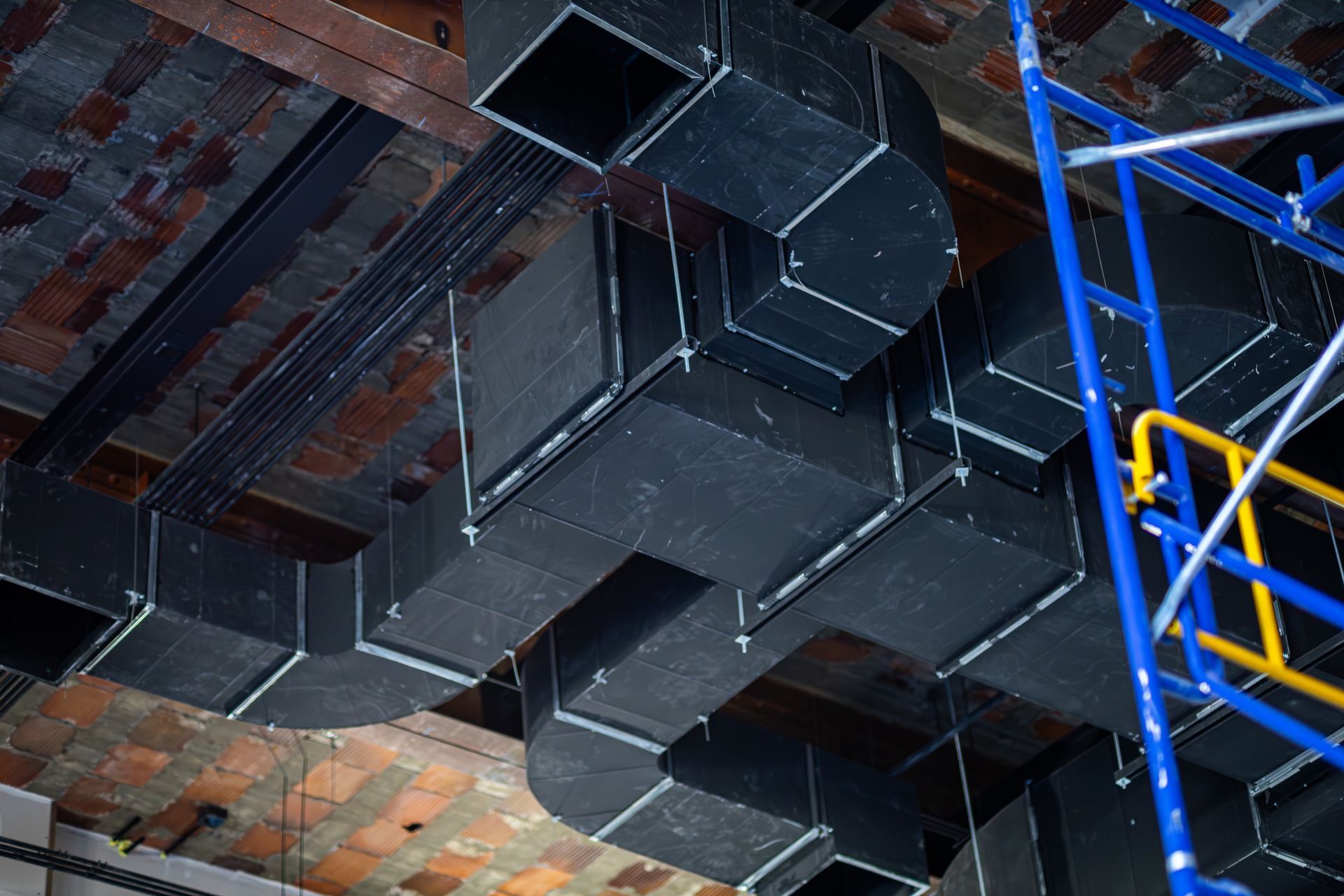 Black HVAC ductwork installed on a brick ceiling, with a blue scaffolding on the right.