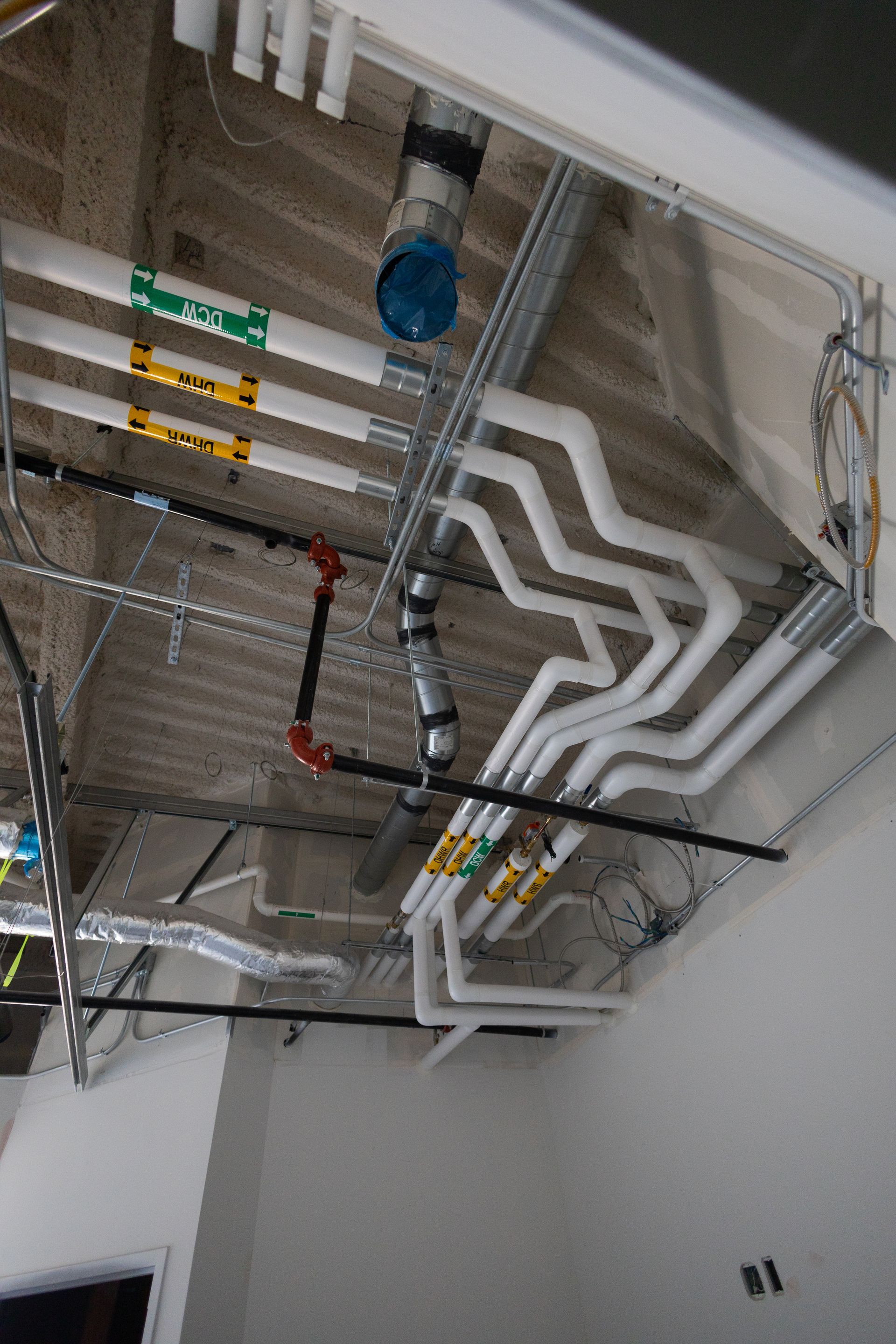 Ceiling view: exposed pipes, conduits, and ventilation in a construction setting, white and silver pipes with colorful labels.