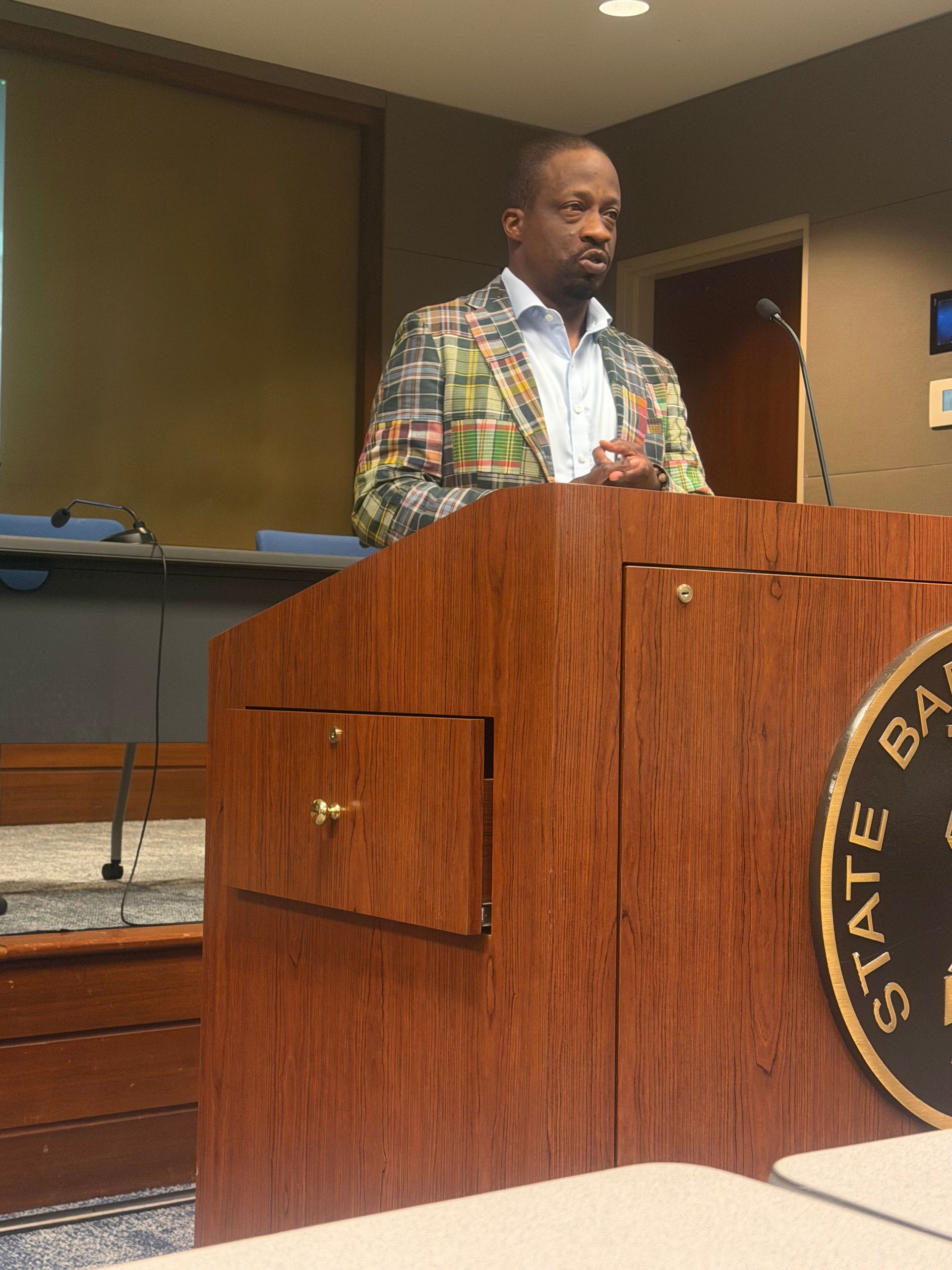 Man speaking at a wooden podium in a courtroom or hearing room, wearing a plaid jacket.