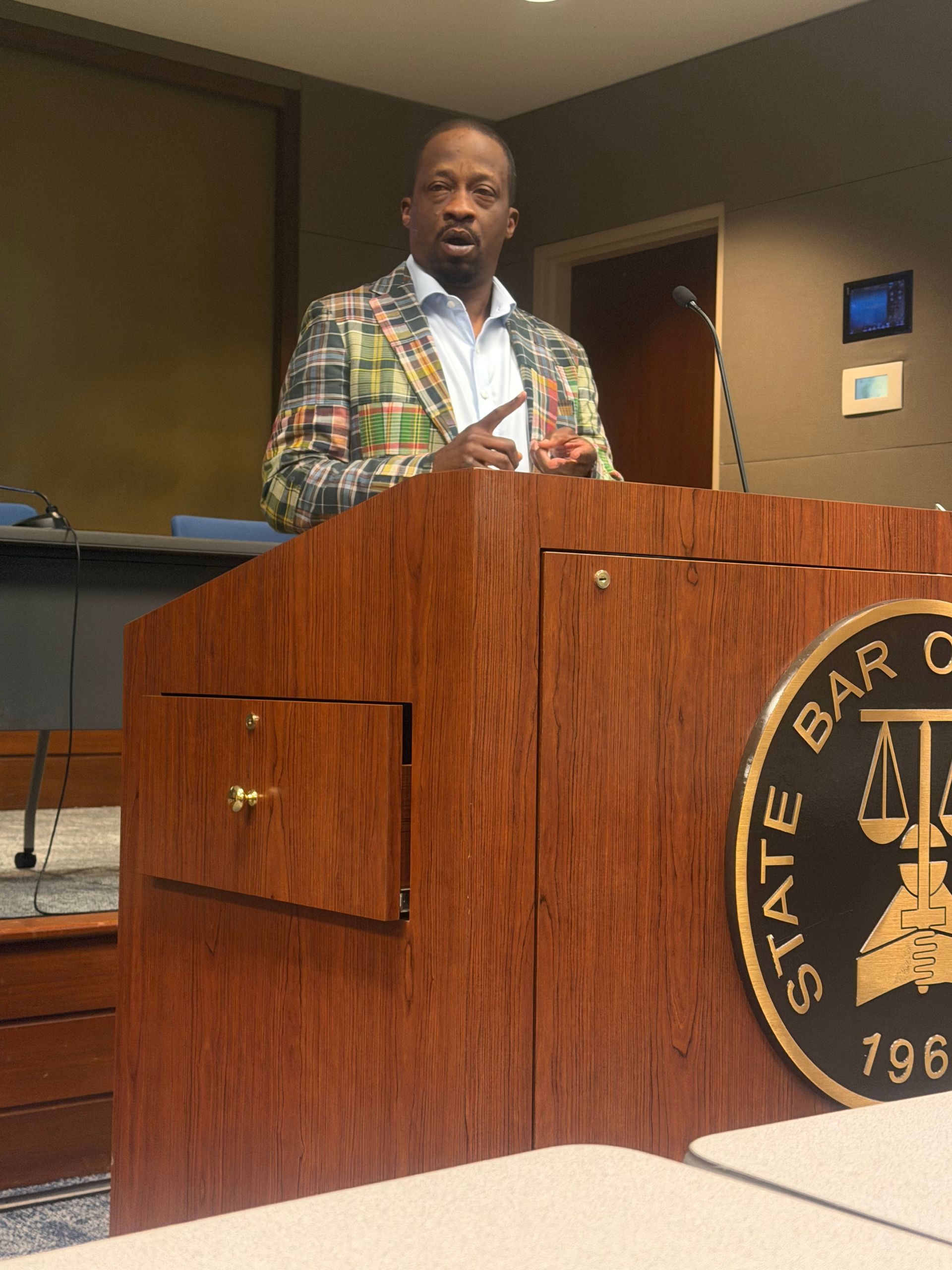 Man speaking at a wooden podium in a courtroom, wearing a colorful plaid jacket.