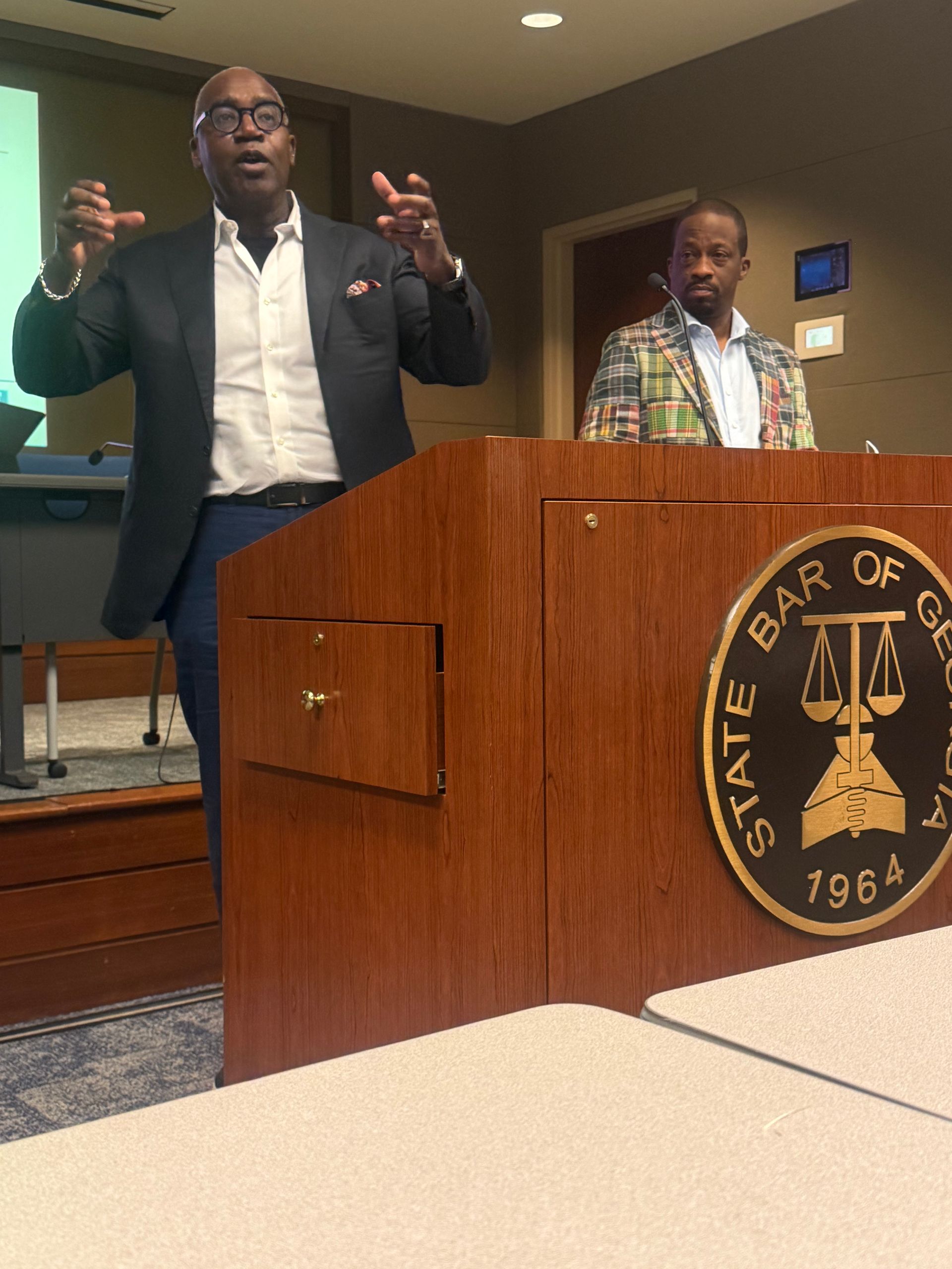 Two speakers at a podium in a courtroom, one gesturing while speaking, with a state bar emblem visible.